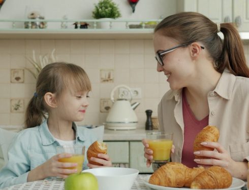 Young woman and adorable little girl are eating croissants and drinking juice talking at breakfast at home, modern interior is visible in background.