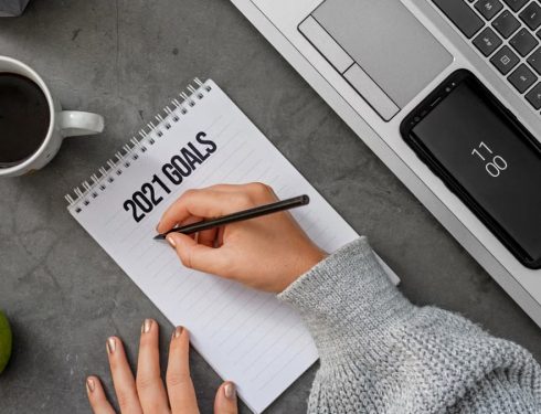 Top view crop anonymous female in sweater writing down goals for 2021 year in notebook while sitting at desk with laptop and cup of coffee