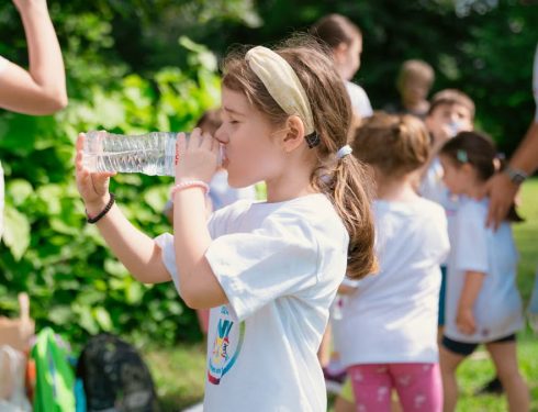 Young girl drinking water during sunny outdoor activity with friends in a park setting.